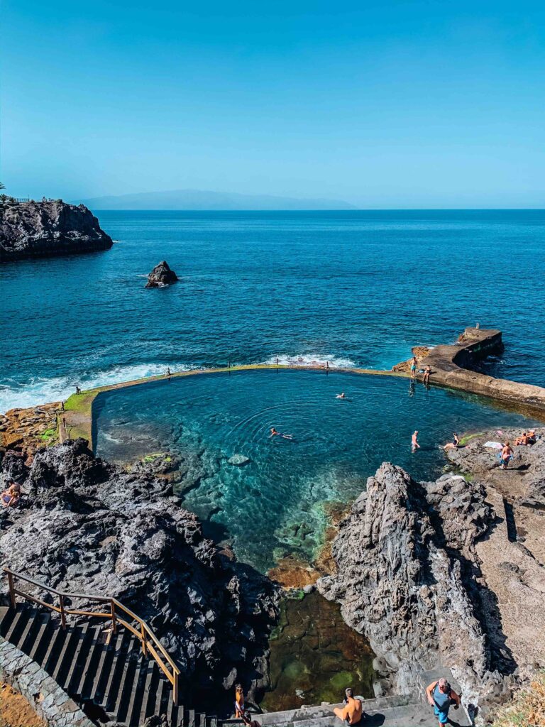11 UNIQUE Boutique Hotels in Tenerife 50 A natural rock pool surrounded by volcanic rocks, with several people swimming and relaxing. Stairs descend from boutique hotels in Tenerife to the pool, while the ocean extends to the horizon under a clear blue sky.