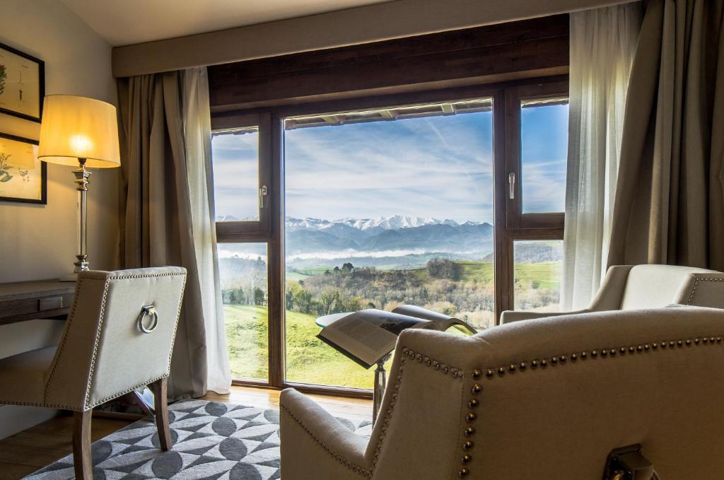 A cozy room with two beige armchairs and a desk, reminiscent of boutique hotels in Asturias, features a large window offering a scenic view of green hills and snow-capped mountains under a blue sky.