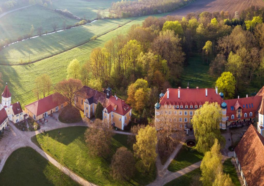 FAIRYTALE Castle Hotels in Bavaria 24 Aerial view of a historic estate reminiscent of castle hotels in Bavaria, with red-roofed buildings surrounded by trees, green fields, and pathways, all bathed in warm sunlight.