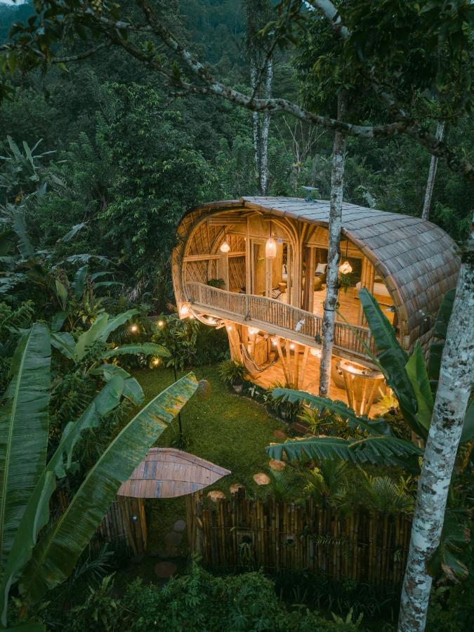 Aerial view of a bamboo treehouse with warm lights, surrounded by lush green tropical plants and trees in a forest setting—an enchanting hideaway that stands among the best family villas in Bali. A small structure and wooden fence grace the garden below.