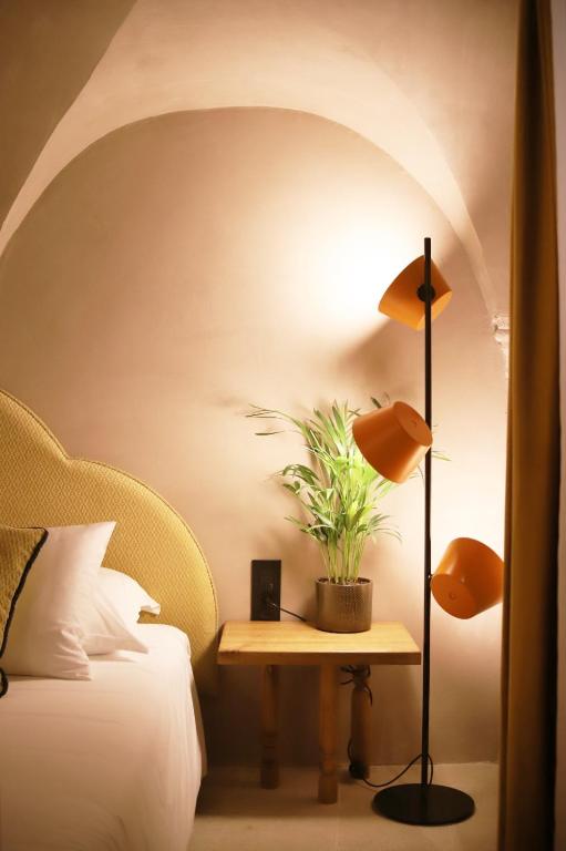 Most Unique Boutique Hotels in Dubrovnik 17 A cozy bedroom corner reminiscent of boutique hotels in Dubrovnik, featuring a yellow cushioned headboard, white bedding, a wooden side table with a potted plant, and a black floor lamp with three orange shades against a curved beige wall.