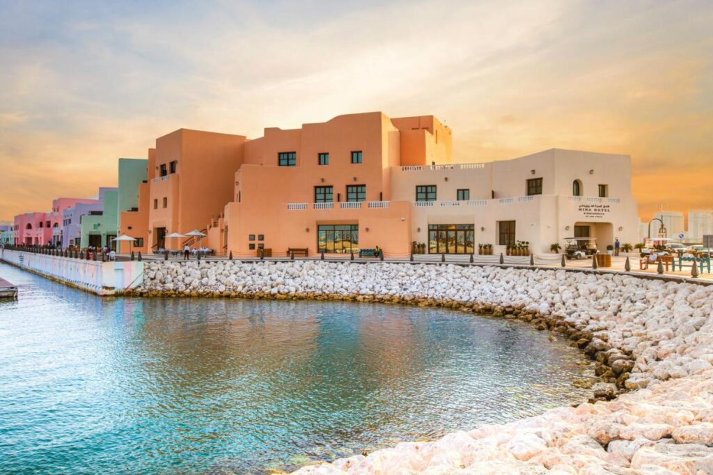 Where to Stay in Doha, Qatar: Transit, City & Beach Hotels 32 Colorful, modern buildings line a waterfront with a rocky shoreline at sunset—an inviting scene that hints at where to stay in Doha for stunning views reflected on calm water beneath a tranquil sky.