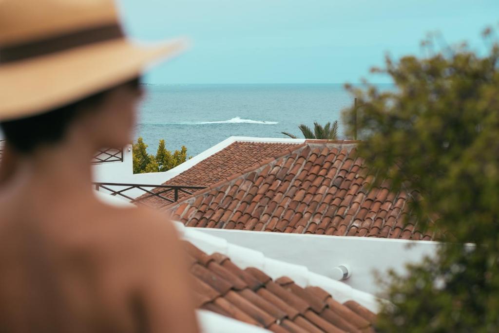 11 UNIQUE Boutique Hotels in Tenerife 33 A person wearing a wide-brimmed hat gazes toward red tile rooftops and the ocean in the distance—perhaps dreaming of Boutique Hotels in Tenerife—while greenery and a clear sky frame the sunlit scene. The blurred foreground draws focus to rooftops and sea.