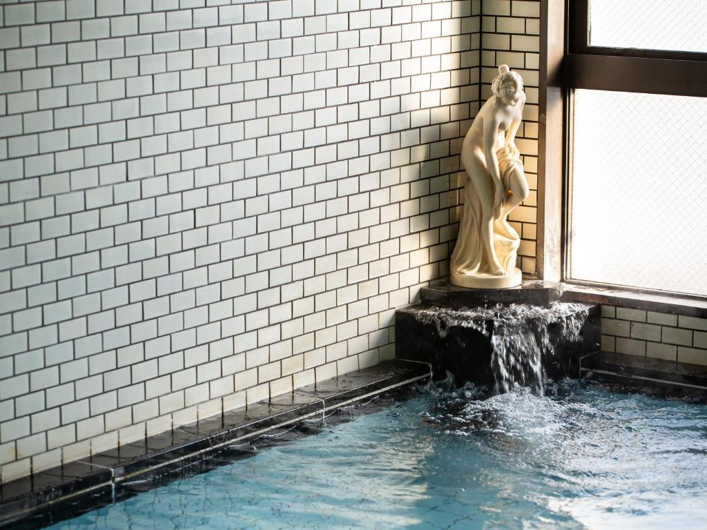 8 BEST Ryokans in Osaka 19 Indoor hot spring bath with white tiled walls, a statue of a woman in the corner, and water flowing from the statue’s base into the pool—reminiscent of serene ryokans in Osaka—with sunlight streaming in from a window.