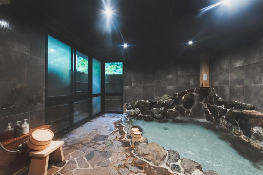 8 BEST Ryokans in Osaka 33 A serene indoor Japanese onsen, often found in ryokans in Osaka, features stone flooring, a natural rock hot spring bath, wooden stools and buckets, and bottles of soap by frosted glass windows. Soft lighting creates a calm, relaxing atmosphere.
