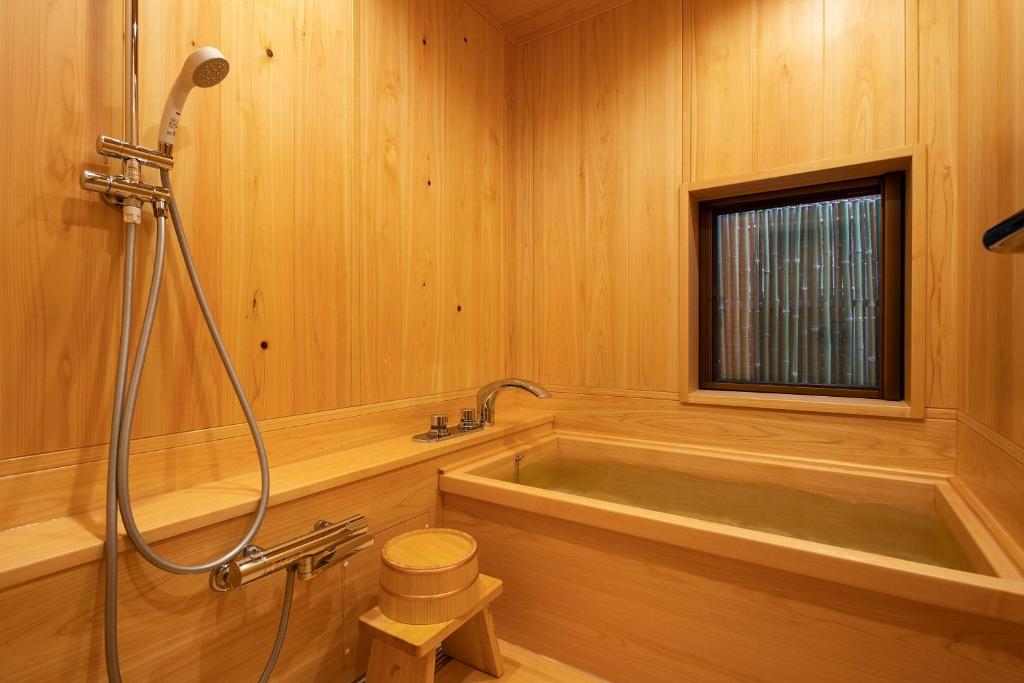 7 UNIQUE Ryokans in Gion Kyoto 30 A traditional Japanese wooden bathroom like those found in ryokans in Gion Kyoto, featuring a deep soaking tub, handheld shower, small wooden stool, and a window with horizontal blinds, all surrounded by light wood paneling.