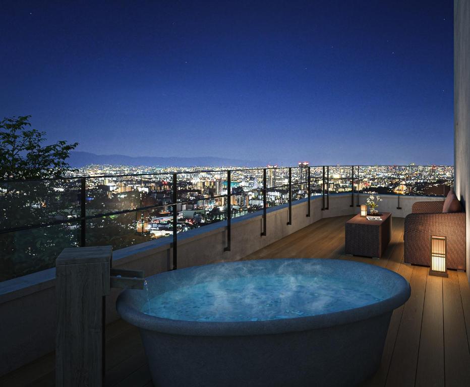 8 BEST Ryokans in Osaka 27 A rooftop terrace at night with a round outdoor hot tub, wooden deck, wicker chairs, and soft lantern lighting evokes the luxury of ryokans in Osaka, overlooking a brightly lit cityscape under a clear, starry sky.