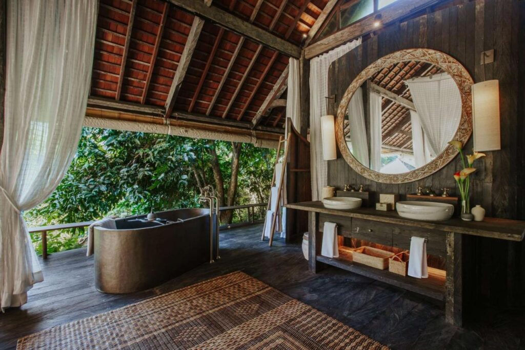 13 Most UNIQUE Places to Stay in Ubud 17 Luxurious open-air bathroom with a large metal bathtub, double sinks, circular mirror, wooden vanity, wicker baskets, and sheer curtains—one of the unique places to stay in Ubud—overlooking lush green trees through wide open windows.