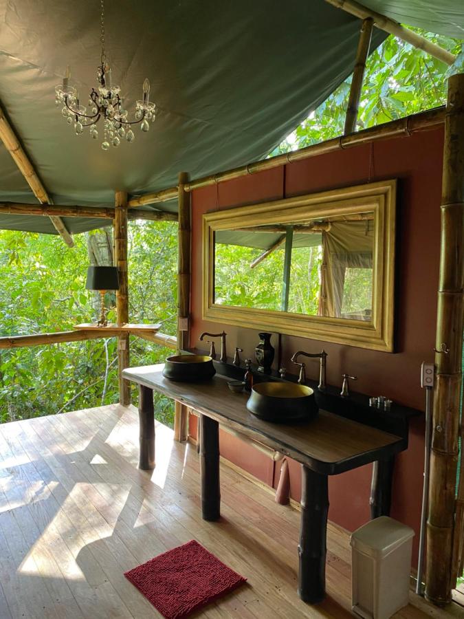 Unique Hotels in Costa Rica: Top Stays 13 A rustic bathroom in one of the unique hotels in Costa Rica features bamboo and wood decor, a double sink, large framed mirror, chandelier, lush green foliage through open walls, and a red rug on the wooden floor.