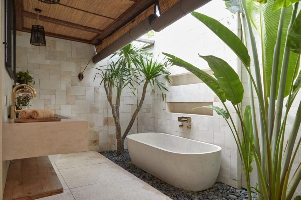 Modern outdoor bathroom with a white freestanding tub, stone tile walls, tall green tropical plants, and natural light—a feature found in some of the best family villas in Bali.