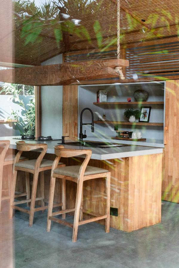 A modern kitchen with wooden cabinets, a kitchen island with three wooden barstools, a black faucet, and open shelves with plants—just like those found in the best family villas in Bali—where greenery is beautifully reflected in the glass.