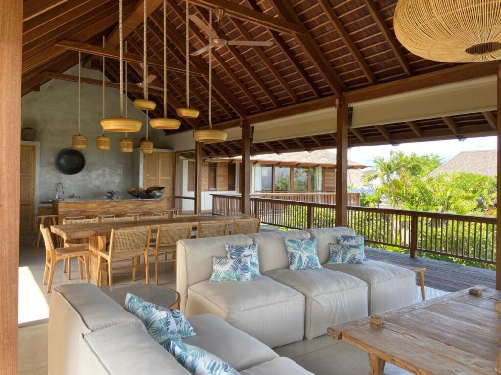 A spacious open-air living area in one of the best family villas in Bali, featuring a white sectional sofa, blue-patterned pillows, a wooden dining table with chairs, modern hanging lights, and views of lush greenery through wooden railings.