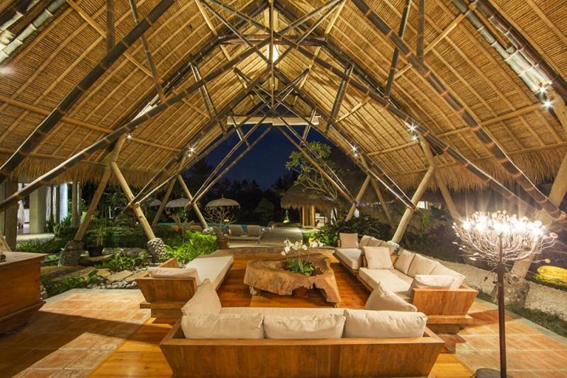 Open-air lounge at one of the best family villas in Bali, featuring a lofty thatched bamboo roof, wooden furniture with cream cushions, a rustic coffee table, and lush greenery beyond, warmly illuminated in the evening.
