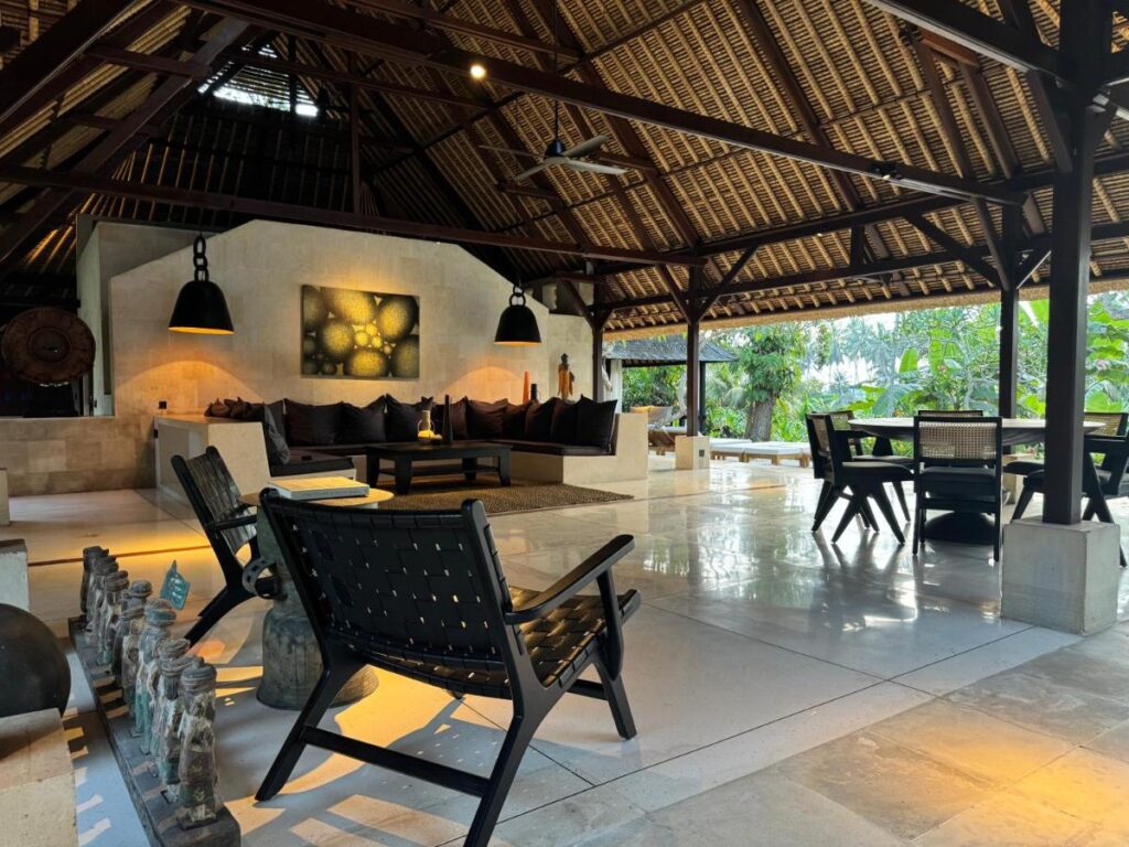Open-air lounge with woven chairs, a large sofa, and a dining table under a high, thatched ceiling. Warm lighting, modern decor, and lush greenery create a relaxed vibe—perfect for those seeking the best family villas in Bali.