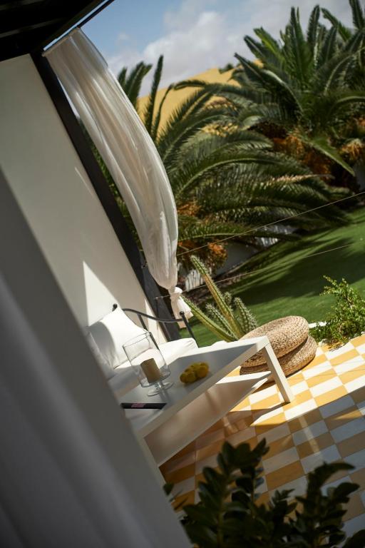 Adults Only Hotels in Fuerteventura 20 A cozy outdoor patio at one of the adults only hotels in Fuerteventura features a white bench, glass of coffee, and lemons on a table. Sunlight filters through a sheer curtain, casting shadows on yellow-checked tiles and lush palm trees.