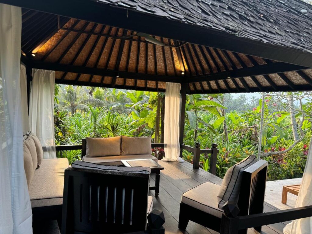 A cozy outdoor seating area with cushioned chairs and a sofa under a wooden gazebo, surrounded by lush tropical plants. Soft natural light filters in, creating the tranquil atmosphere found at the best family villas in Bali.