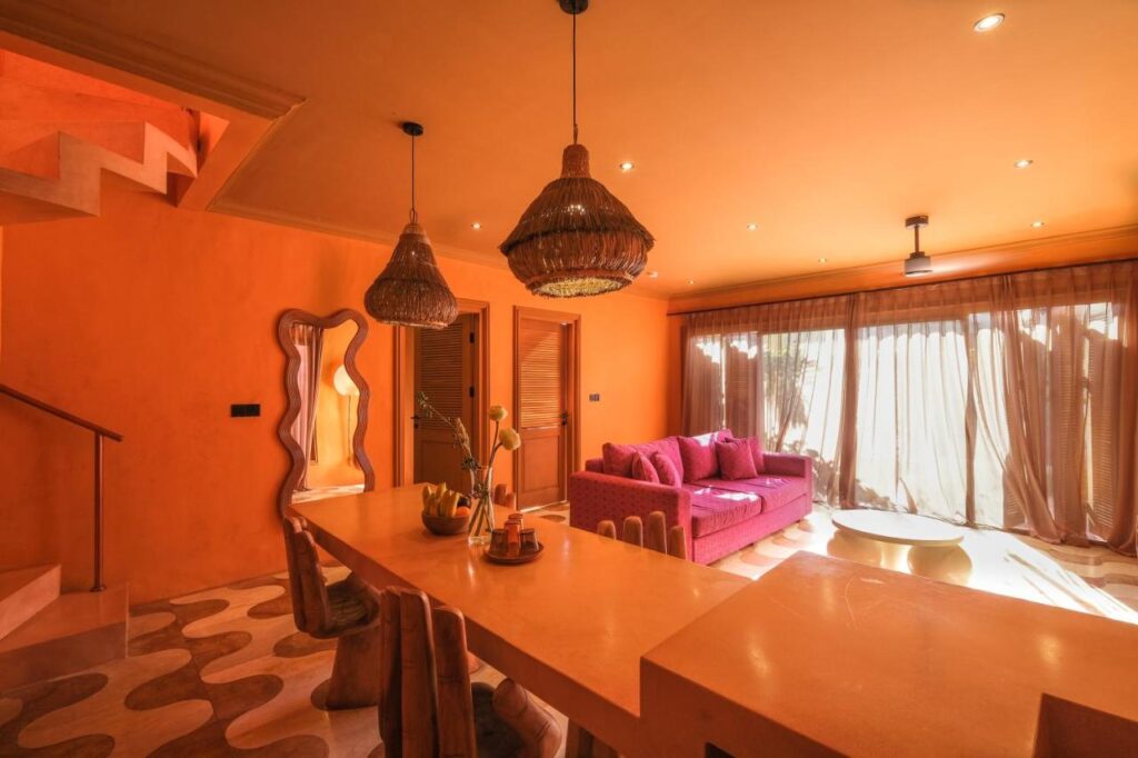 14 UNIQUE Boutique Hotels in Canggu, Bali 49 A warm, orange-toned living space inspired by boutique hotels in Canggu features a long dining table, wooden chairs, woven pendant lights, a bold pink sofa, patterned rug, and large windows with sheer curtains letting in sunlight.