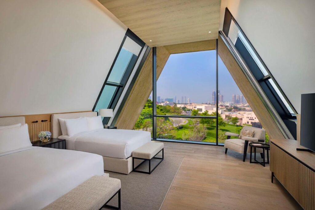 Where to Stay in Doha, Qatar: Transit, City & Beach Hotels 8 A modern hotel room with two double beds, light wood floors, large floor-to-ceiling windows in a triangular frame, cityscape views—an ideal choice for travelers searching where to stay in Doha. Minimalistic decor, white walls, TV, and chair complete the space.