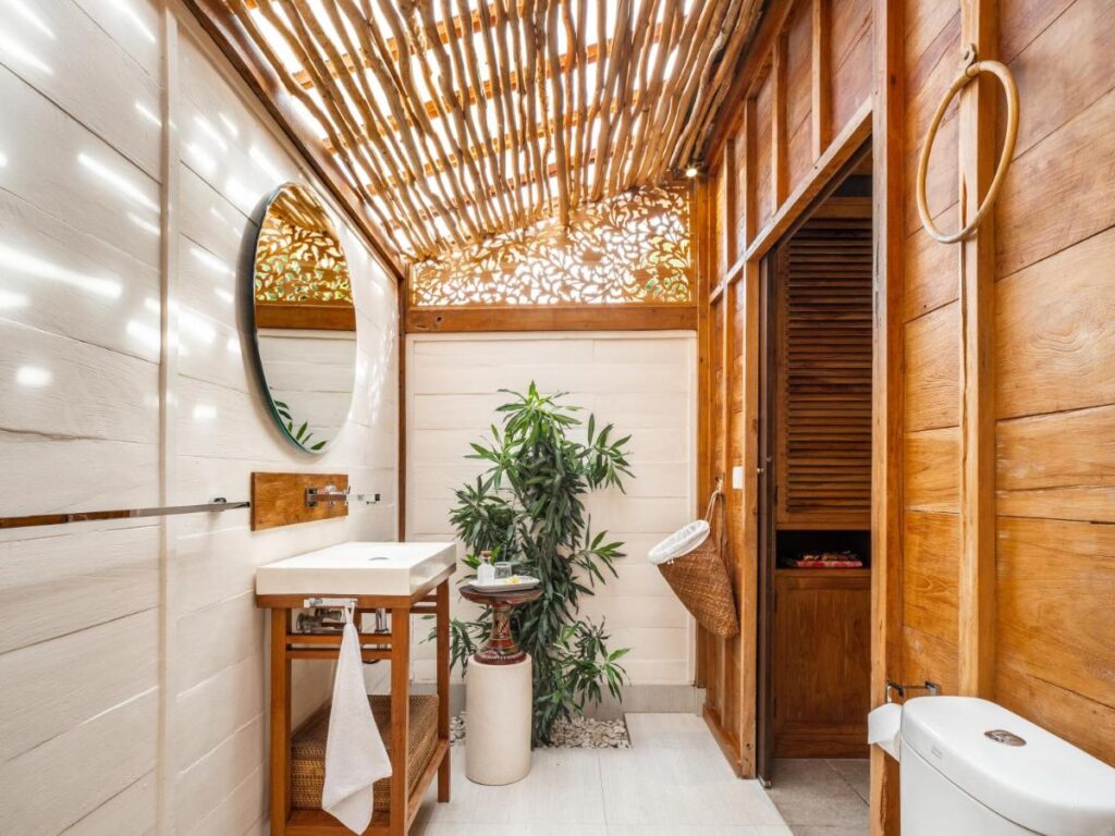14 UNIQUE Boutique Hotels in Canggu, Bali 45 A modern bathroom, inspired by boutique hotels in Canggu, features wooden and white walls, a round mirror above the sink, a potted plant near the center, sleek wooden shelving, and a ceiling of wooden slats welcoming natural light.