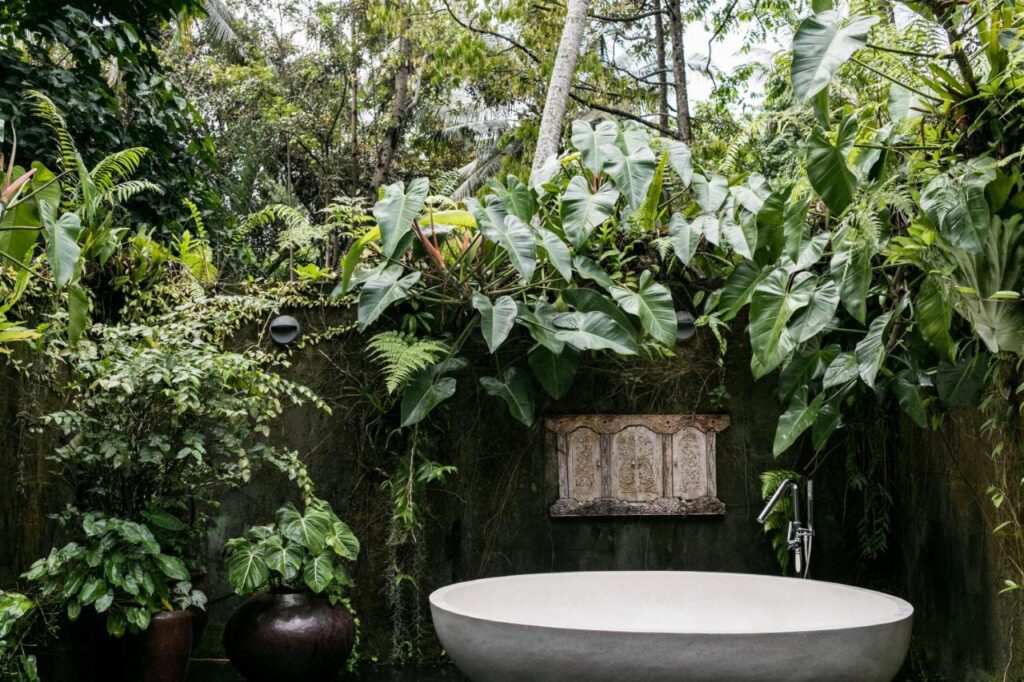 13 Most UNIQUE Places to Stay in Ubud 8 An outdoor bathroom with a large stone bathtub surrounded by lush green tropical plants, a decorative wall feature, and a dark ceramic pot—one of the unique places to stay in Ubud for a serene, nature-inspired setting.