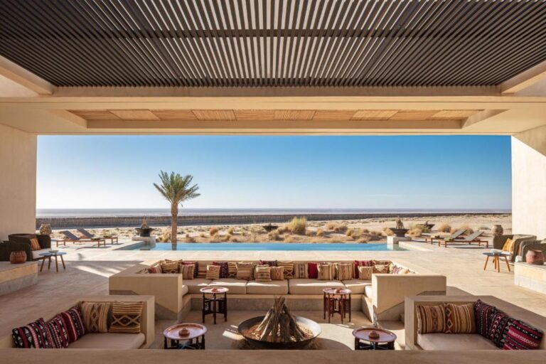 Open-air lounge with patterned cushions around a fire pit, overlooking a pool, palm trees, sun loungers, and a sandy beach—experience the charm of boutique hotels in Tunisia against a backdrop of sea and clear blue sky.