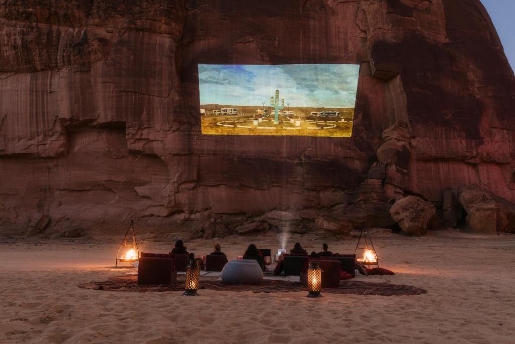 9 Best Hotels in Saudi Arabia: Unique Stays Across the Country 25 A group of people watches a movie projected onto a large rock wall in a desert, seated on sofas and cushions with lanterns and rugs on the sand at sunset—an outdoor cinema experience inspired by the Best Hotels in Saudi Arabia.