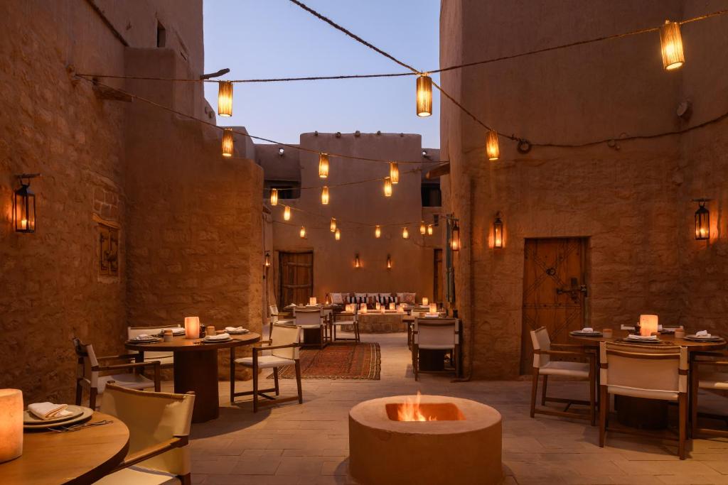 9 Best Hotels in Saudi Arabia: Unique Stays Across the Country 21 An outdoor restaurant at one of the Best Hotels in Saudi Arabia features stone walls, wooden tables and chairs, and warm lantern lights. A central fire pit glows, creating a cozy and inviting atmosphere at dusk.