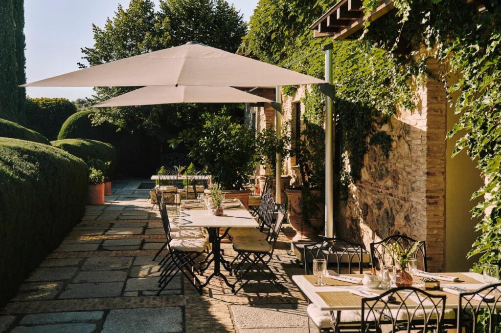 25 Weekend Trips from Madrid Max. 2h Away 89 An outdoor dining area with neatly set tables, black chairs, and a large white umbrella, surrounded by lush greenery and ivy-covered stone walls—an inviting spot often discovered on weekend trips from Madrid.