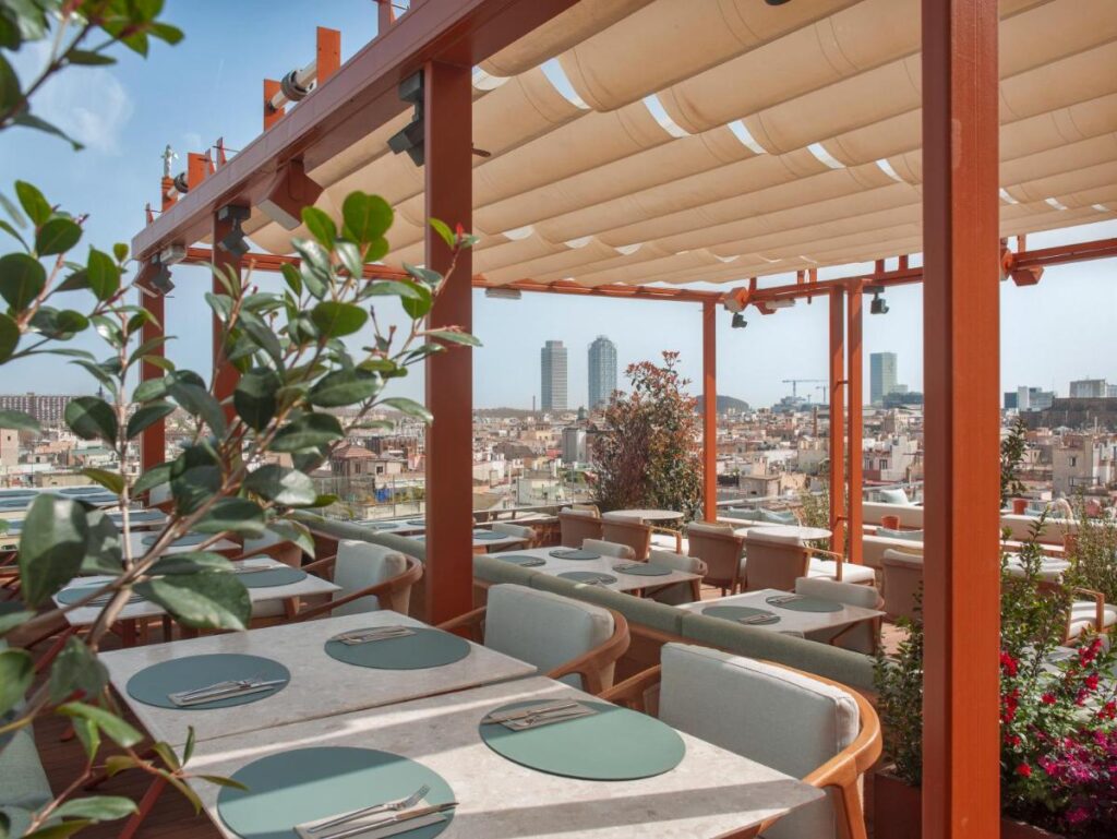 Top Barcelona Hotels with Rooftop Pool 24 Outdoor rooftop restaurant with neatly set tables and chairs under a pergola with cream fabric shades, overlooking a cityscape of tall buildings and greenery—ideal for guests seeking the ambiance found in Barcelona hotels with rooftop pool views.