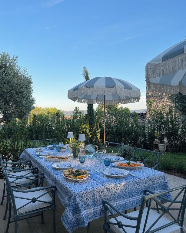 11 Unique Places to Stay in Provence 29 Outdoor dining table set with plates of food, glasses, and a blue-patterned tablecloth. Surrounded by chairs, greenery, and a blue-and-white striped umbrella under a clear sky—capturing the charm of unique places to stay in Provence.