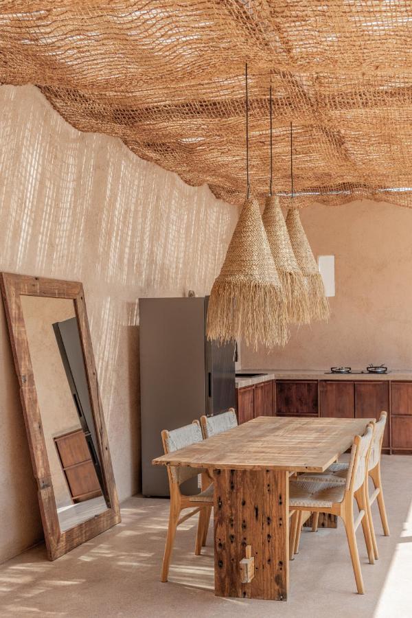 A rustic dining area in one of the best family villas in Bali, featuring a wooden table, four chairs, and a large leaning mirror. Woven straw pendant lights hang from a netted ceiling as natural light plays on the beige walls.