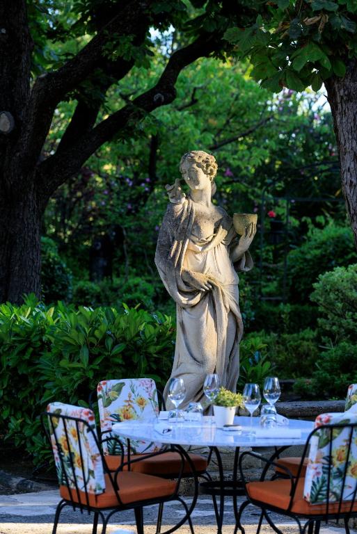 11 Unique Places to Stay in Provence 35 A stone statue of a woman holding a cup stands in a lush garden. In the foreground, a round table set with wine glasses and floral chairs creates an inviting outdoor dining scene, evoking the charm of unique places to stay in Provence.