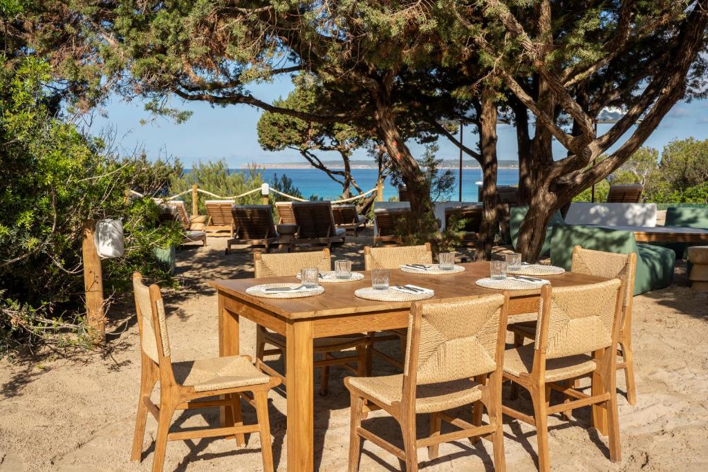A wooden outdoor dining table with six chairs sits on sandy ground beneath trees, overlooking a scenic seaside view with lounge chairs in the background—an inviting setting often found at boutique hotels in Formentera.