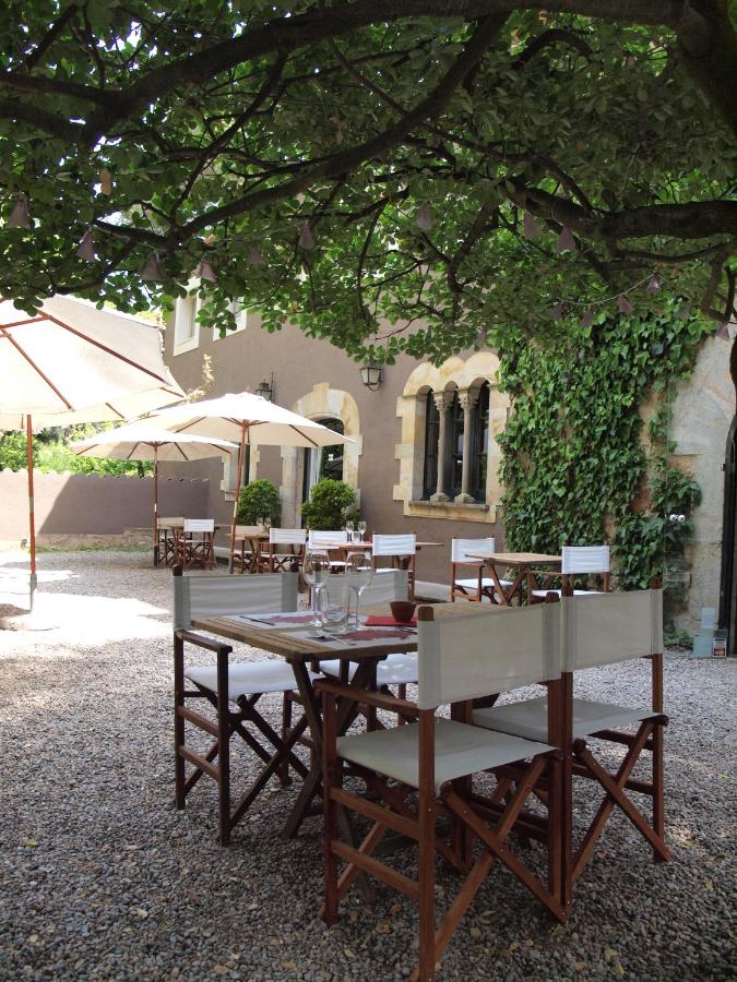 Outdoor dining area with wooden chairs and tables set on gravel, shaded by a large tree, with stone buildings and ivy in the background—an inviting scene found at many boutique hotels in Costa Brava. Sun umbrellas offer extra shade.