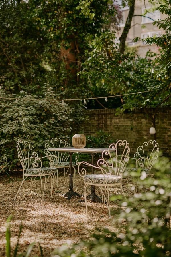 15 Best Boutique Hotels in London, UK 3 An outdoor garden seating area with a round table and four ornate metal chairs, surrounded by greenery and trees—a setting reminiscent of the charming courtyards found at the best boutique hotels London offers. A lantern graces the table, with string lights above.