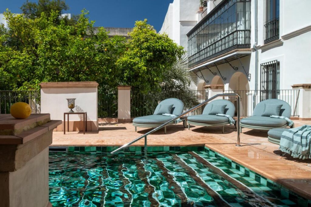 10 UNIQUE Hotels with Pool in Seville 35 A small outdoor pool with green and white tiles is surrounded by cushioned round loungers, leafy trees, and a white building with iron balconies under a clear blue sky—perfect for those seeking hotels with pool in Seville.