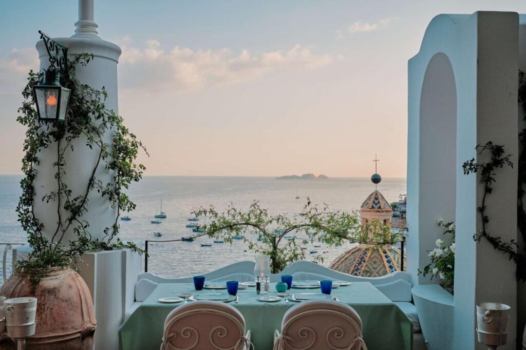 Boutique Hotels on the Amalfi Coast 2 A romantic outdoor dining table overlooks the sea, with blue glasses and plates set for four—a scene reminiscent of boutique hotels on the Amalfi Coast. White columns and flowering vines frame boats and a colorful domed church at sunset.