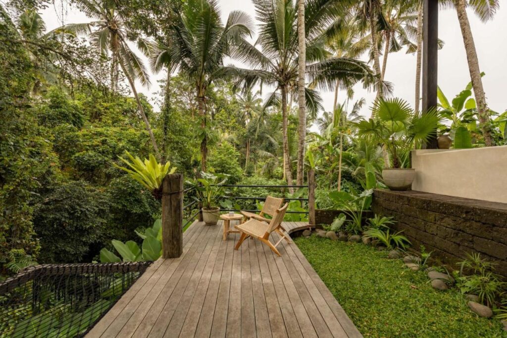 A wooden deck overlooking lush tropical greenery with palm trees, a rope net, two wooden chairs, and potted plants—creating a relaxing outdoor seating area in a jungle setting often found at the best family villas in Bali.