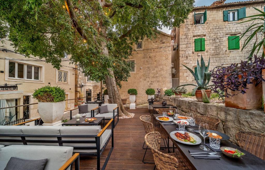 8 TOP Boutique Hotels in Split 34 Outdoor restaurant patio with modern seating, set tables, and food, surrounded by potted plants and large trees. Historic stone buildings with green shutters recall the charm found in boutique hotels in Split, framing this cozy, inviting space.