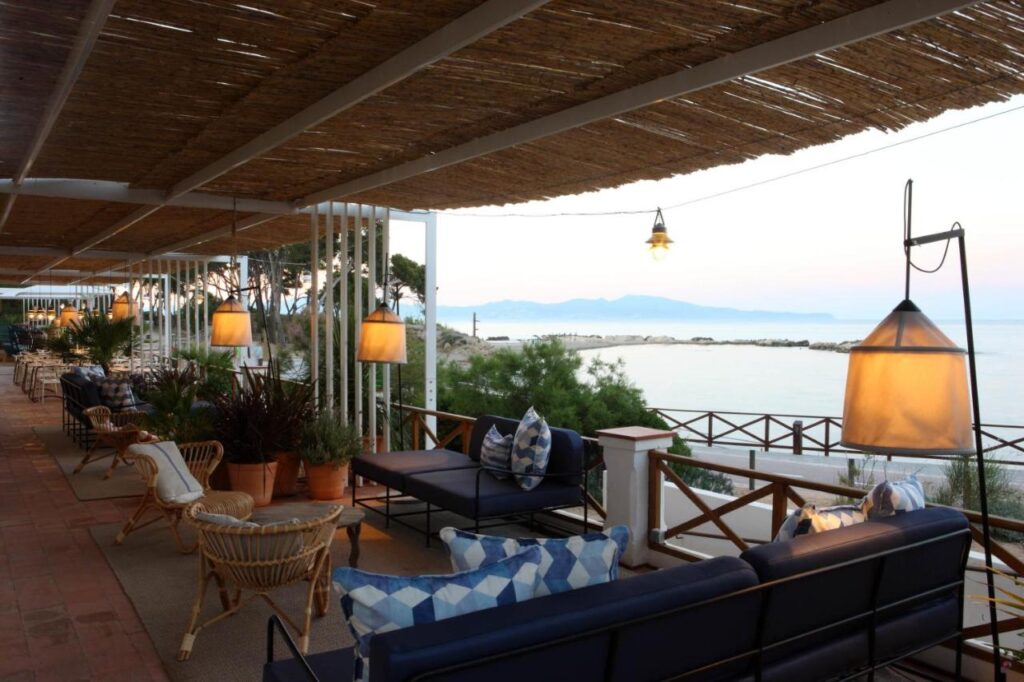 Outdoor seating area with cozy sofas, wicker chairs, and warm hanging lights overlooking a calm sea and distant mountains at sunset—shaded by a wooden pergola, this relaxed coastal spot evokes the charm of boutique hotels in Costa Brava.