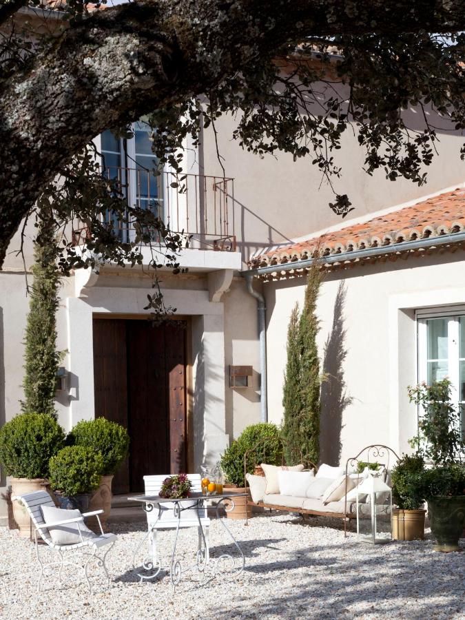 25 Weekend Trips from Madrid Max. 2h Away 4 A sunlit Mediterranean patio, perfect for weekend trips from Madrid, features a white metal table and chairs, potted plants, cushioned bench, and a large tree casting shade before a stucco house with tall windows and a wooden door.