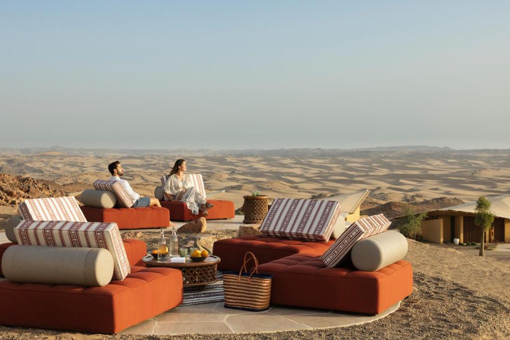 9 Best Hotels in Saudi Arabia: Unique Stays Across the Country 28 Two people relax on patterned cushions atop modern red outdoor sofas, enjoying a scenic desert landscape near one of the Best Hotels in Saudi Arabia; a picnic basket and refreshments are arranged nearby under the clear sky.