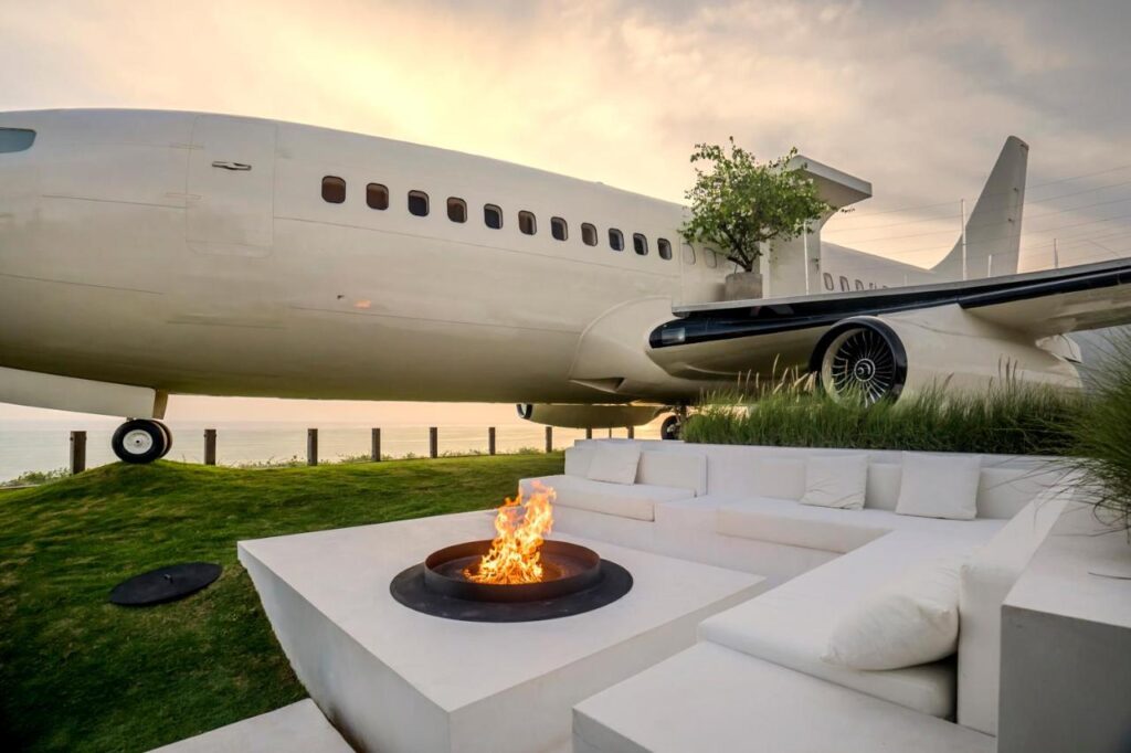 A large, white airplane is repurposed as a building near a firepit with modern white outdoor seating and green grass—a unique spot reminiscent of the best family villas in Bali, complete with a tree growing through the wing under a bright, cloudy sky.