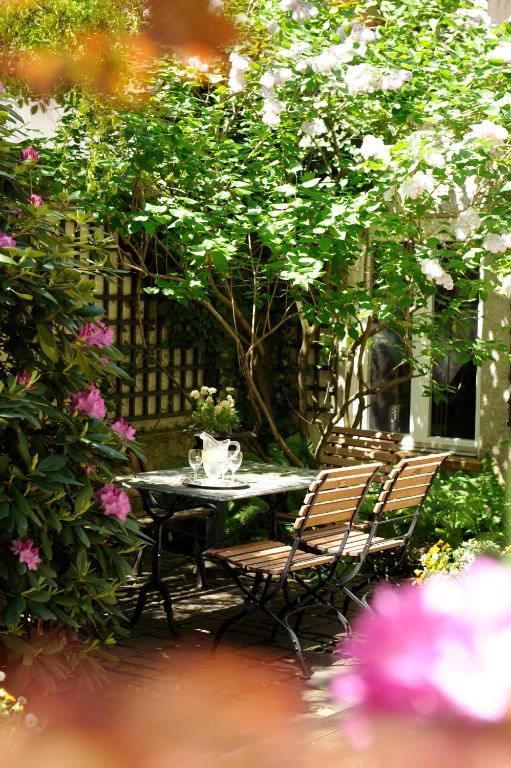 Authentic Boutique Hotels in Munich 19 A sunlit garden patio at one of the boutique hotels in Munich features a small table and two wooden chairs surrounded by lush greenery and blooming flowers. A glass pitcher and cups sit on the table, creating a peaceful, inviting atmosphere.