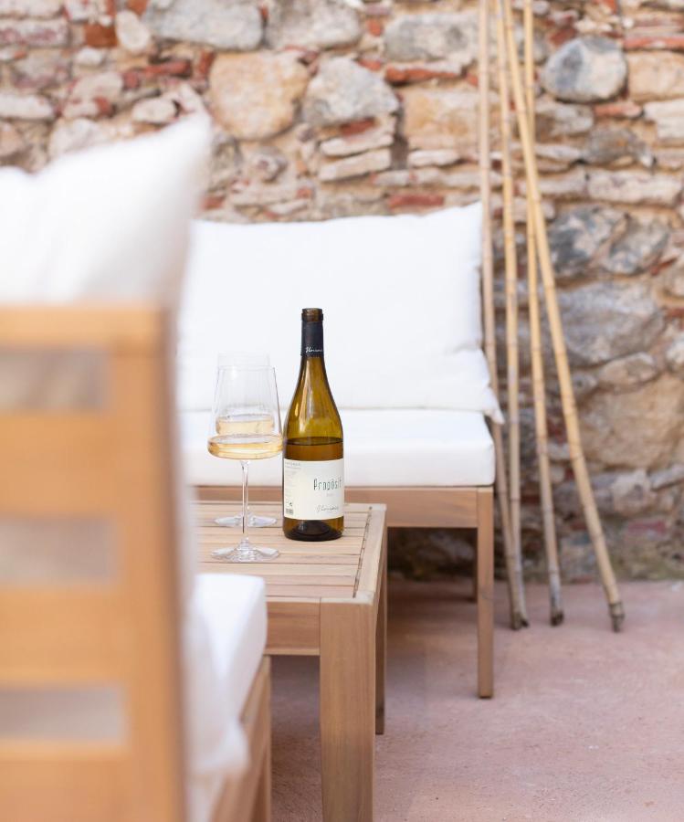 A bottle of white wine and a filled glass rest on a wooden table between cushioned outdoor chairs, set against a stone wall with bamboo sticks—evoking the relaxed elegance found at boutique hotels in Costa Brava.