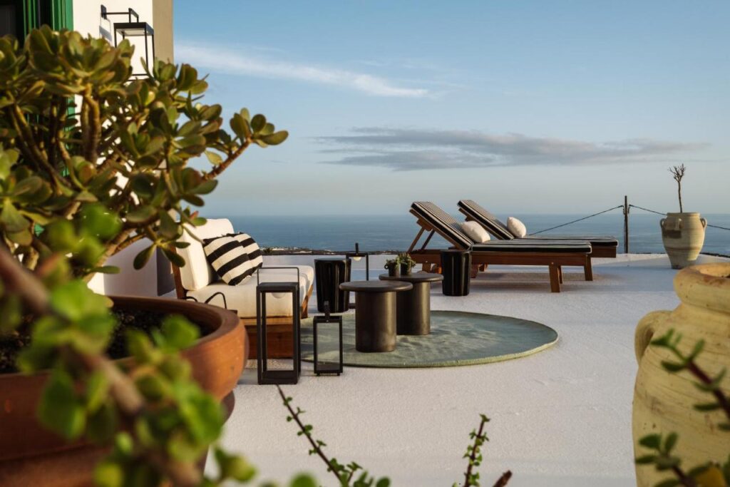 Unique Adults Only Hotels in Lanzarote 2 A sunny terrace at one of the Adults Only Hotels in Lanzarote, with lounge chairs, side tables, potted plants, and lanterns, overlooks a calm ocean under a clear sky for a relaxing seaside atmosphere.