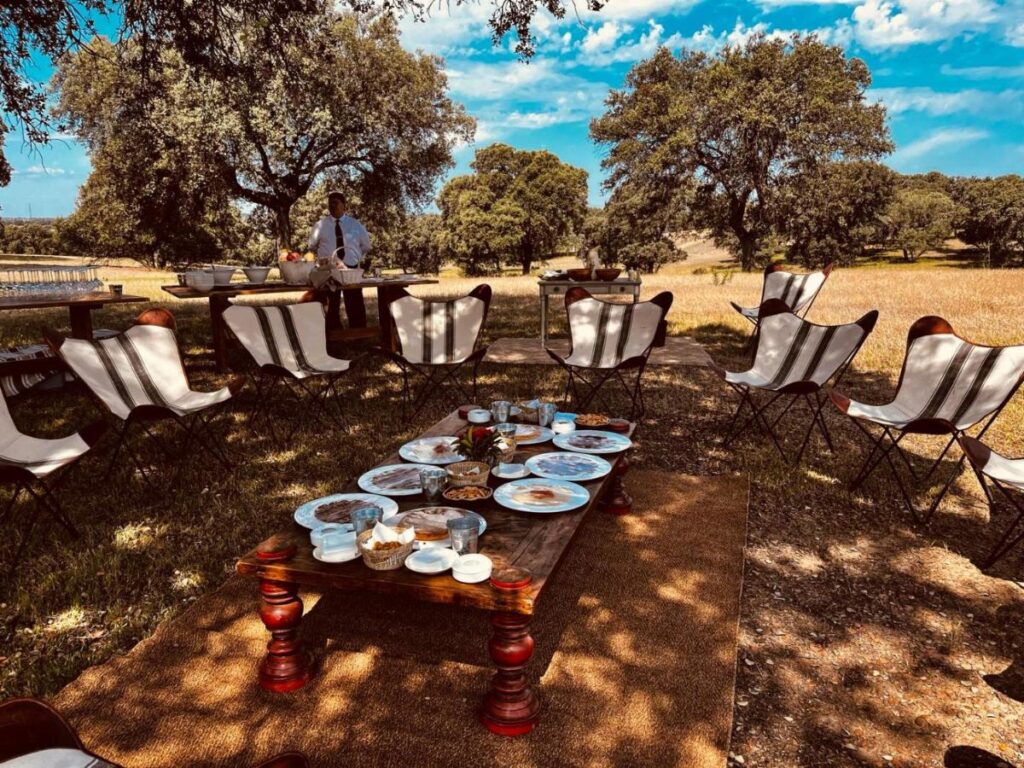 25 Weekend Trips from Madrid Max. 2h Away 6 A rustic outdoor setting perfect for weekend trips from Madrid, with a low wooden table surrounded by white butterfly chairs. Used plates and cups rest on the table as a person stands near buffet tables under large trees in a sunny grassy field.