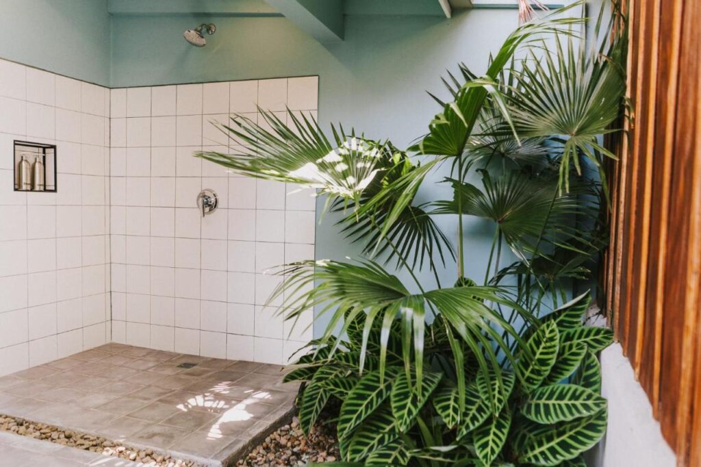 Unique Hotels in Costa Rica: Top Stays 17 An outdoor shower with white tiled walls and a metal showerhead, surrounded by lush green tropical plants. Pebbles line the floor next to the shower, and natural light streams in from above—hallmarks of unique hotels in Costa Rica.