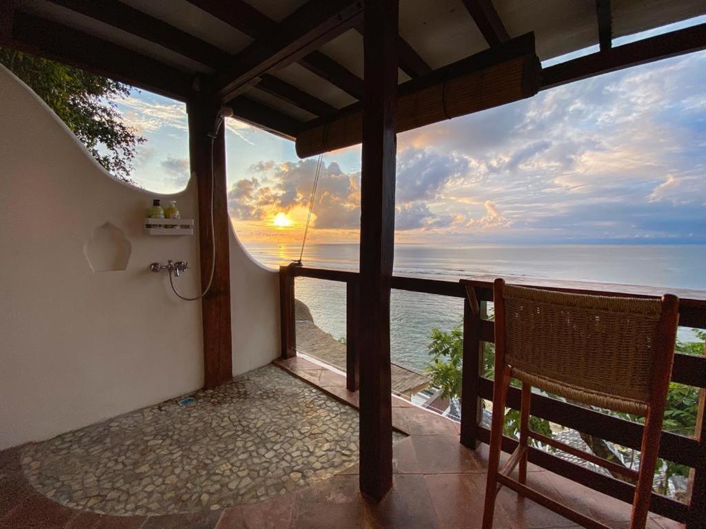 A serene outdoor patio with a wooden chair overlooks the ocean at sunset, framed by a stucco wall and wooden beams—bottles on a shelf and a pebble floor complete this peaceful, idyllic scene reminiscent of the best family villas in Bali.