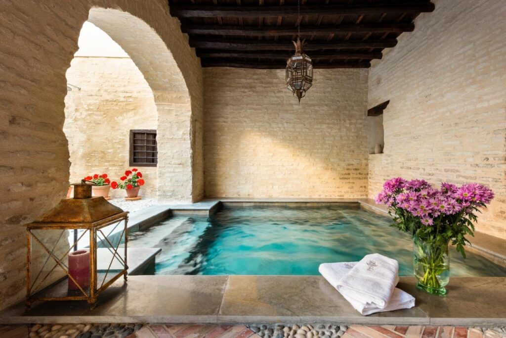 10 UNIQUE Hotels with Pool in Seville 30 A cozy indoor spa pool in the style of hotels with pool in Seville, featuring stone walls, wooden ceiling beams, steps into the water, a lantern with a candle, pink flowers in a vase, and neatly folded towels on the edge.